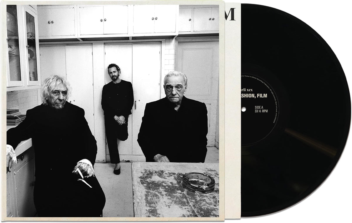A black-and-white vinyl record partially pulled from its sleeve, showing a monochrome photograph of three men in a room. The record label text is partially visible on the black disc.